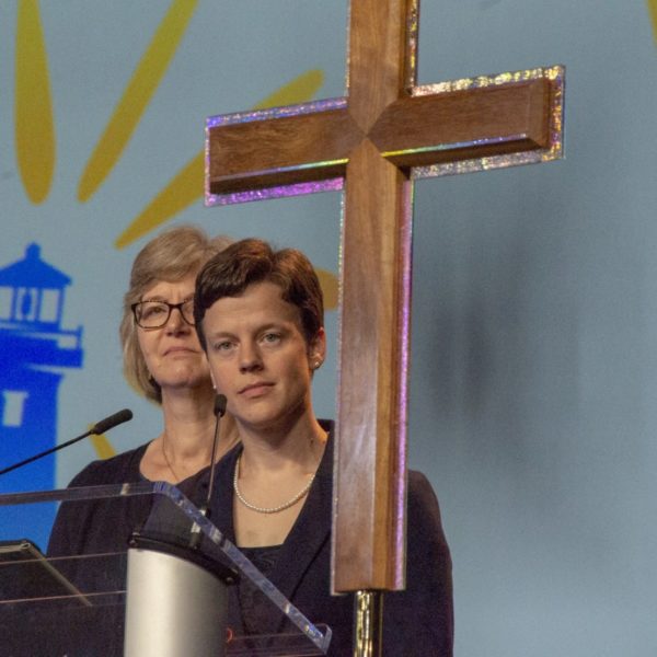 a person stands on stage behind a microphone and large, wooden cross
