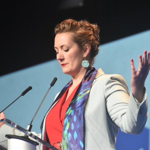 a pastor stands behind a podium with her arms stretched out and head bowed